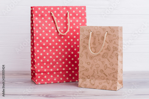 Two natural paper shopping bags. Red and brown paper gift bags with printing on light wooden background. Business, retail, sale and commerce concept.