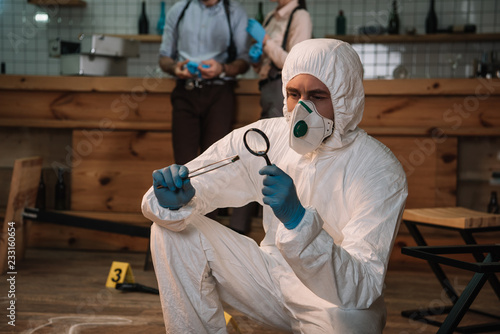 focused forensic investigator examining evidence with magnifying glass at crime scene with colleagues working behind