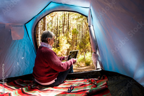 Mature adult retired woman sitting inside a tent in free wild camping alone in the forest using a technology internet connected tablet to organize the travel for digital nomad work