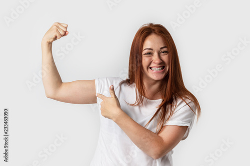 Portrait smiling woman in white t-shirt , isolated on gray background. Showing biceps muscle having lifted arm with clenched fist
