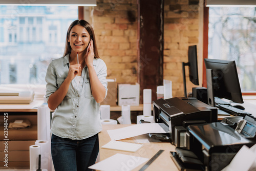 Portrait of charming woman in blue shirt having phone conversation while standing near office desk with printer. She is looking away and smiling