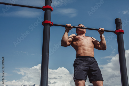 Caucasian male athlete with fit torso and strong hands doing chin ups outdoor. Handsome blonde sportsman pulls up on stadium horizontal bar. Chin up bar exercises.