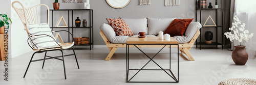Wood and metal coffee table with candles and tea cup in bright living room interior with grey lounge with cushions and chair, metal racks with decor and plant in vase in real photo
