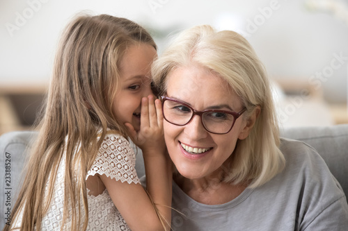 Cute little granddaughter whispering in ear telling secret to understanding smiling grandma, kid girl secretly talking to granny having fun gossiping, trust in grandmother and grandchild relations