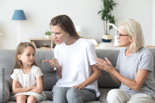 Angry young mother scolding child daughter while old grandmother interfering when strict mom lecturing little girl, three generations disagreements affecting upbringing kid, family conflicts concept