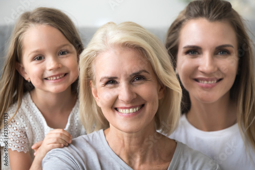 Smiling senior grandmother looking at camera with young daughter and kid granddaughter at background, older grandma in three 3 women happy loving family, older elderly generation concept, portrait