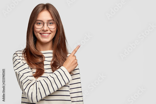 Waist up shot of pretty Caucasian woman with cheerful expression, points with index finger at blank copy space, dressed in striped sweater, shows free space at upper right corner for your promotion