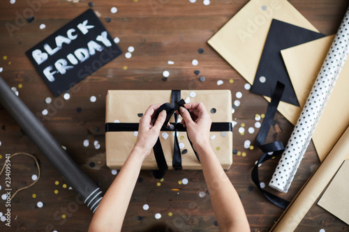 Top view of female hands tying ribbon around wrapped present box over wooden table with decorative stuff and card Black Friday