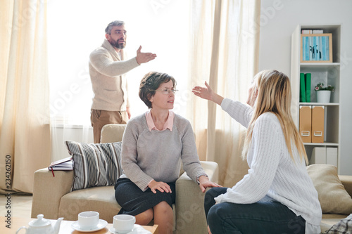 Frowning family psychologist in glasses sitting in armchair and listening to quarrel of couple while asking young lady to stop conflict, emotional couple gesturing hands and blaming each other for