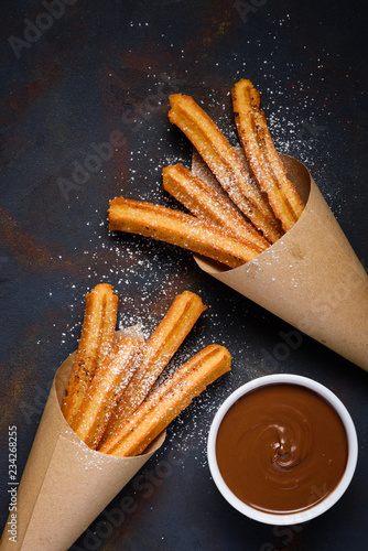 Traditional churros sticks in paper bag with sugar powder cinnamon and bowl of chocolate dip on dark table top view