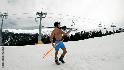 Naked nude male in shorts, helmet, goggles and boots standing on slope under ski lift