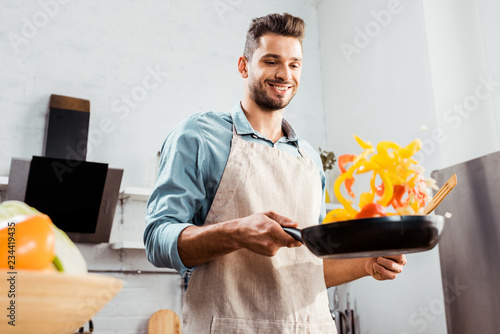 low angle view of smiling young man in apron holding frying pan with vegetables
