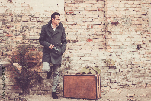 Portrait of thoughtful vintage styled man with leather suitcase leaning on brick wall.