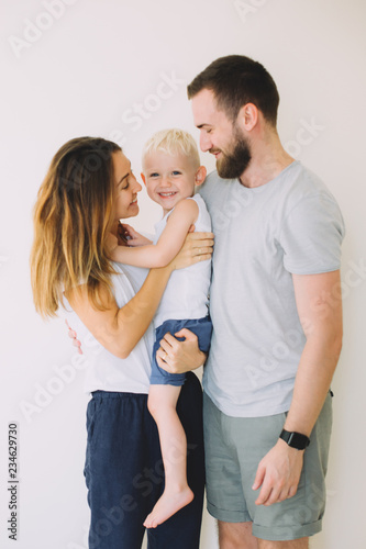 Happy young family with little son standing together in casuals near the empty wall - indoors