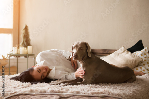 Happy young woman in a pajamas with dog weimaraner sits on a bed at home. Winter or christmas weekend concept.