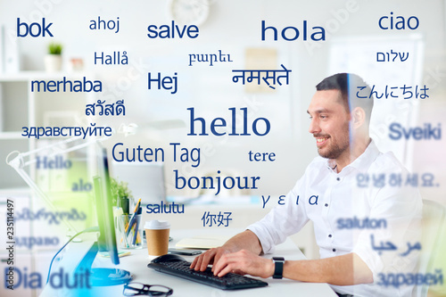 translation, business, and technology concept - smiling male translator or businessman typing on computer keyboard at office over greeting words in different foreign languages