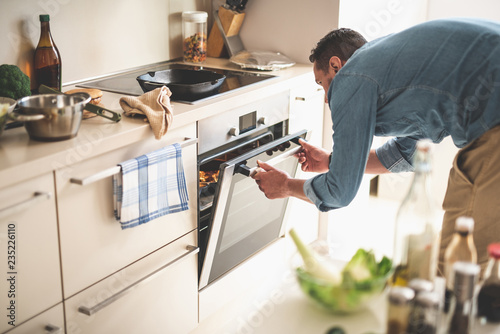 Portrait of gentleman in denim shirt closing door of oven while looking at frying pan with beef steak