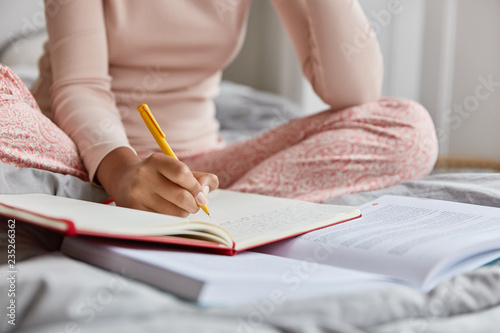 Cropped image of unrecognizable woman in nightclothes, writes down information in notepad, rewrites topic from textbook, poses at bed alone, has nice handwriting. Close up shot, focus on writing