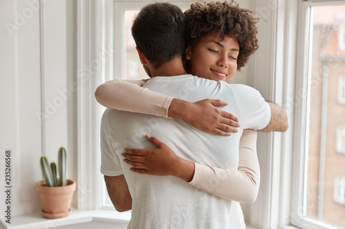 Pleased dark skinned young woman gives warm hug to her boyfriend, being pleased, pose near window, have romantic relationship, stand in cozy room. Husband and wife feel pleased and togetherness