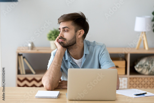 Tired male student or worker sit at home office desk look in distance having sleep deprivation, lazy millennial man distracted from work feel lazy lack motivation, thinking of dull monotonous job