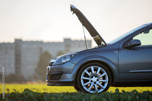 Side view detail of car with open hood on empty gravel field road on blurred apartment building and clear bright sky copy space background. Transportation, vehicles problems and breakdowns concept.