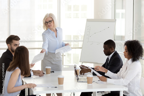 Smiling mature female manager company leader gives handout to diverse employees team at office group meeting, middle aged businesswoman handing paper finanical report to workers at corporate briefing