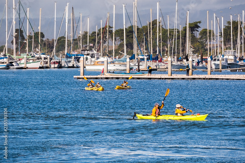 People kayaking in Elkhorn Slough on a sunny day; yachts moored in the marina visible in the background; Moss Landing, California