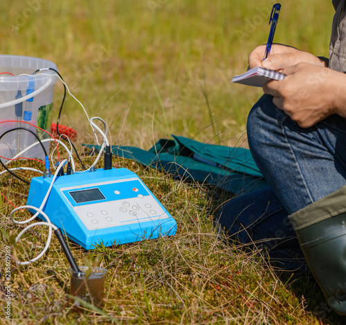 The scientist is writing in his notebook the data from a measuring device. The ecologist and his scientific equipment are in a natural ecosystem.