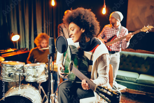 Mixed race woman singing and playing guitar while sitting on chair with legs crossed. In background drummer and bass guitarist.