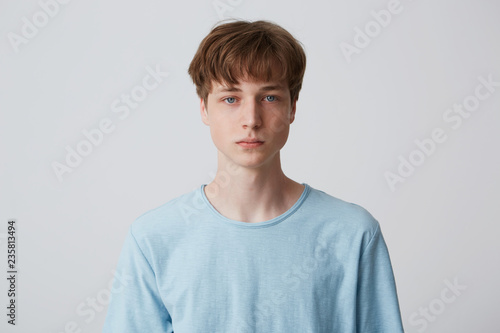 Close up face of a young man without emotions. Beautiful emotionless guy in a blue t-shirt looking to the camera, isolated over white background