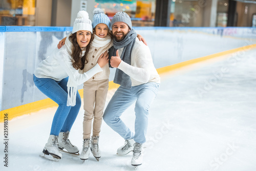 smiling parents and daughter in sweaters looking at camera on skating rink