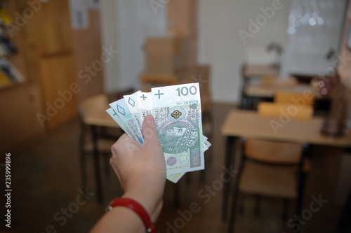 Kobieca ręka wyciągnięta z plikiem banknotów 100 i 50 złotych, w tle puste biuro / wnętrze, z biurkami, szfkami, krzesłami