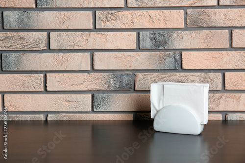 Ceramic napkin holder with paper serviettes on table near brick wall. Space for text