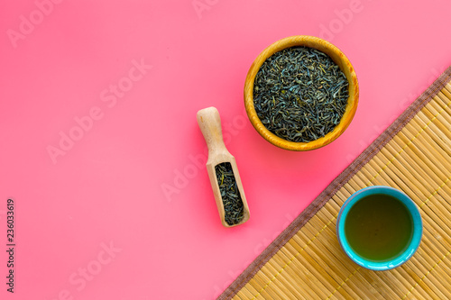 Chinese tea concept. Tea ceremony. Dry tea leaves in bowl and wooden scoop near cup of tea on pink background and bamboo table mat top view copy space