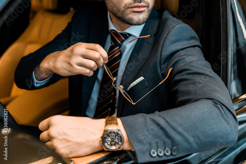 partial view of businessman with luxury watch holding eyeglasses while sitting in car