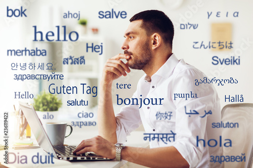 translation, business, and technology concept - male translator or businessman with laptop computer thinking at office over greeting words in different foreign languages