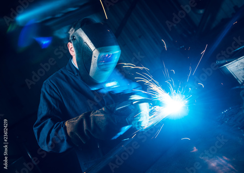 A vibrant action shot of a skilled working metal welder in action, welding metal. Photographed with a slow shutter speed and spark trails. Orange and teal.