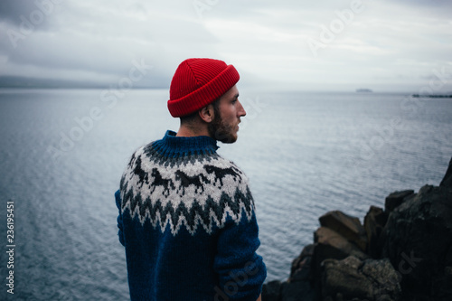Young handsome man with beard wears traditional authentic blue wool knitted sweater with ornaments and red fisherman or sailor beanie hat, looks to horizon, pensative and thoughtful