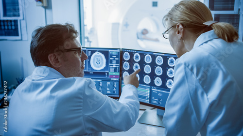 In Control Room Doctor and Radiologist Discuss Diagnosis while Watching Procedure and Monitors Showing Brain Scans Results, In the Background Patient Undergoes MRI or CT Scan Procedure.