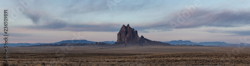 Dramatic panoramic landscape view of a dry desert with a mountain peak in the background during a vibrant cloudy sunrise.Taken at Shiprock, New Mexico, United States.