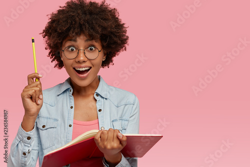 Indoor shot of cheerful dark skinned student keeps pencil in hand, feels happy as gets good idea for writing composition, holds notepad, works on article, looks joyfully isolated over pink studio wall