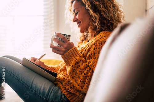 Happy cute lady at home write notes on a diary while drink a cup of tea and rest and relax taking a break. autumn colors and people enjoying home lifestyle writing messages or lists. Blonde curly 