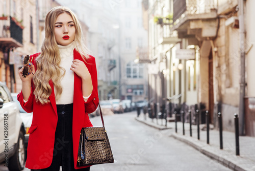 Outdoor fashion portrait of young beautiful fashionable woman wearing white turtleneck, red blazer, holding stylish faux reptile skin handbag, model posing in street of european city. Copy space