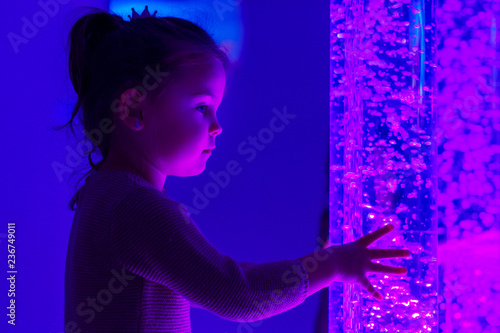 Child in therapy sensory stimulating room, snoezelen. Child interacting with colored lights bubble tube lamp during therapy session.