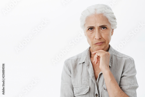 Waist-up shot of concerned serious-looking perplexed elderly woman with grey hair frowning and smirking from dissatisfaction holding hand on chin thinking trying solve troublesome situation