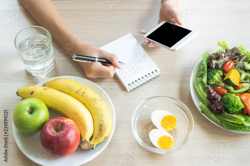Dieting and calories control for wellness. Woman using smartphone calculate calories of food in breakfast during dieting for lose weight program and take notes.