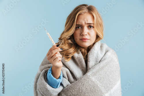 Image of ill upset woman 20s wrapped in blanket holding thermometer, isolated over blue background