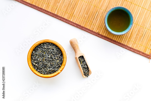 Chinese tea concept. Tea ceremony. Dry tea leaves in bowl and wooden scoop near cup of tea on white background and bamboo table mat top view copy space