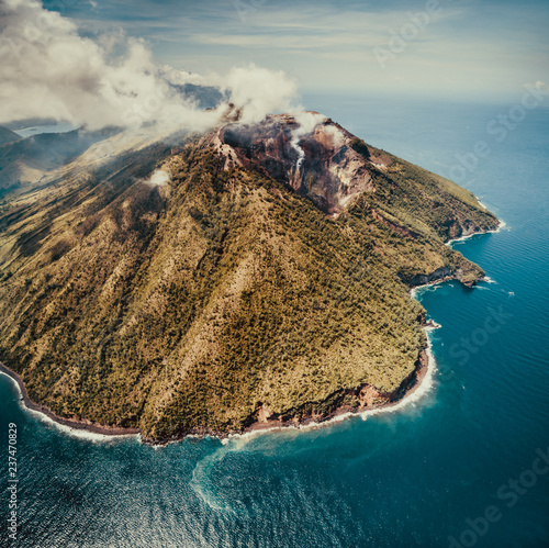 Fog over Indonesia volcano. Aerial drone shot. Amazing overview of Indonesian land surrounded by the ocean. Asian landscape. Active volcano among the calm blue ocean water. Cloudy sky background.
