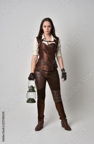 full length portrait of brunette girl wearing brown leather steampunk outfit. standing pose holding a gas lantern on grey studio background.